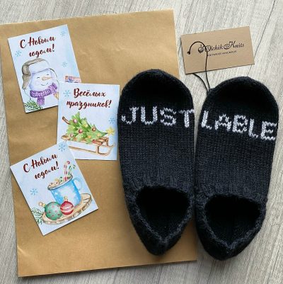 Чёрные вязаные следки с белой вышитой надписью JUST LABLE, рядом новогодние открытки и бирка Olchik Knits на крафтовой бумаге
